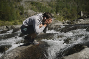 製品を身に着け、山あいの滝のそばで、ハイキング中に顔を洗い、川の水を飲む男性